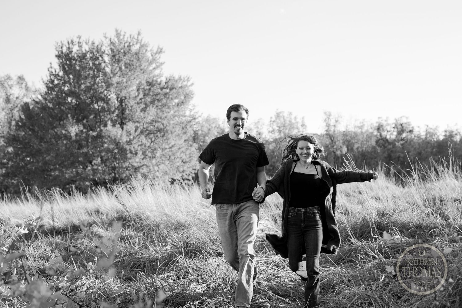 A couple runs joyfully through a field with their dog in a black and white photo.