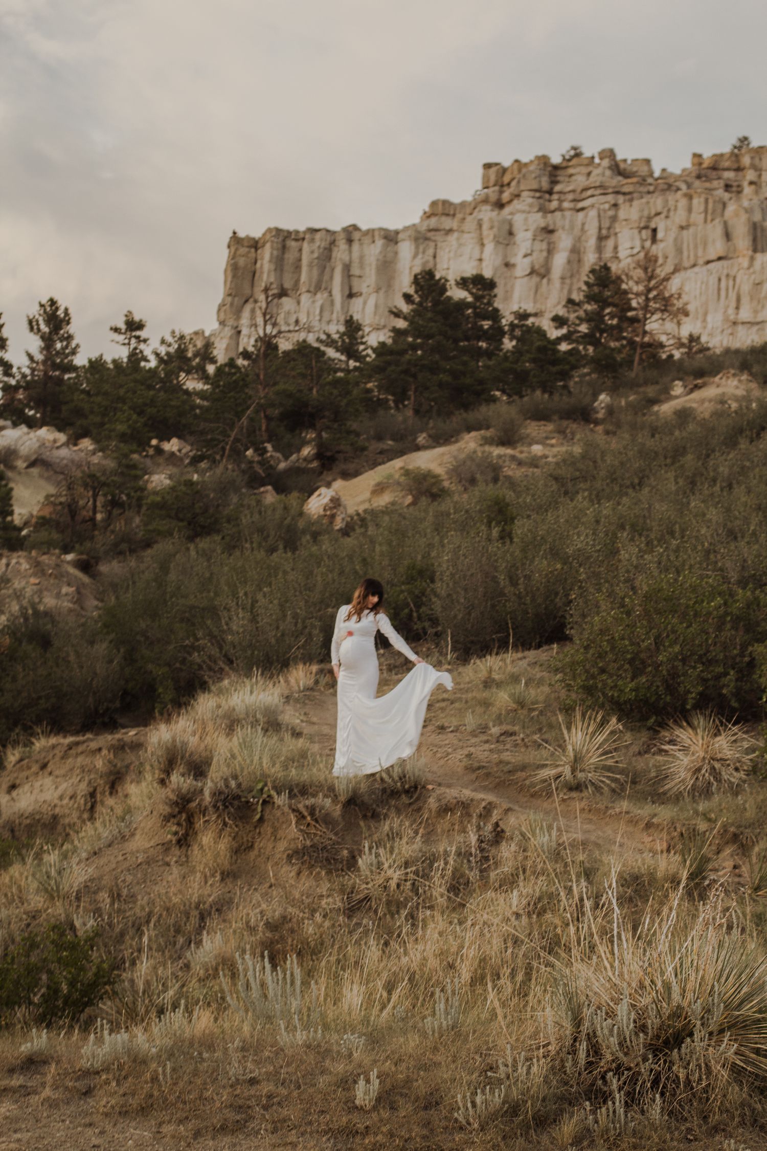 White dress flowing in wind against dramatic rocky cliffs and desert brush landscape.