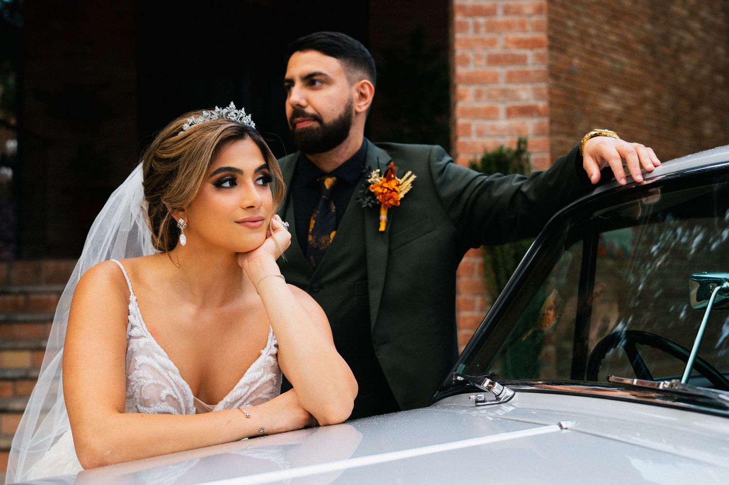 A couple poses with a classic car outside a brick building.