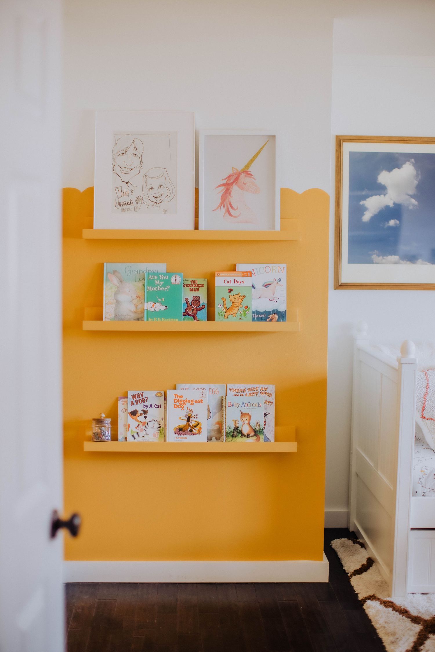 Yellow accent wall with floating shelves displaying children's books and artwork.