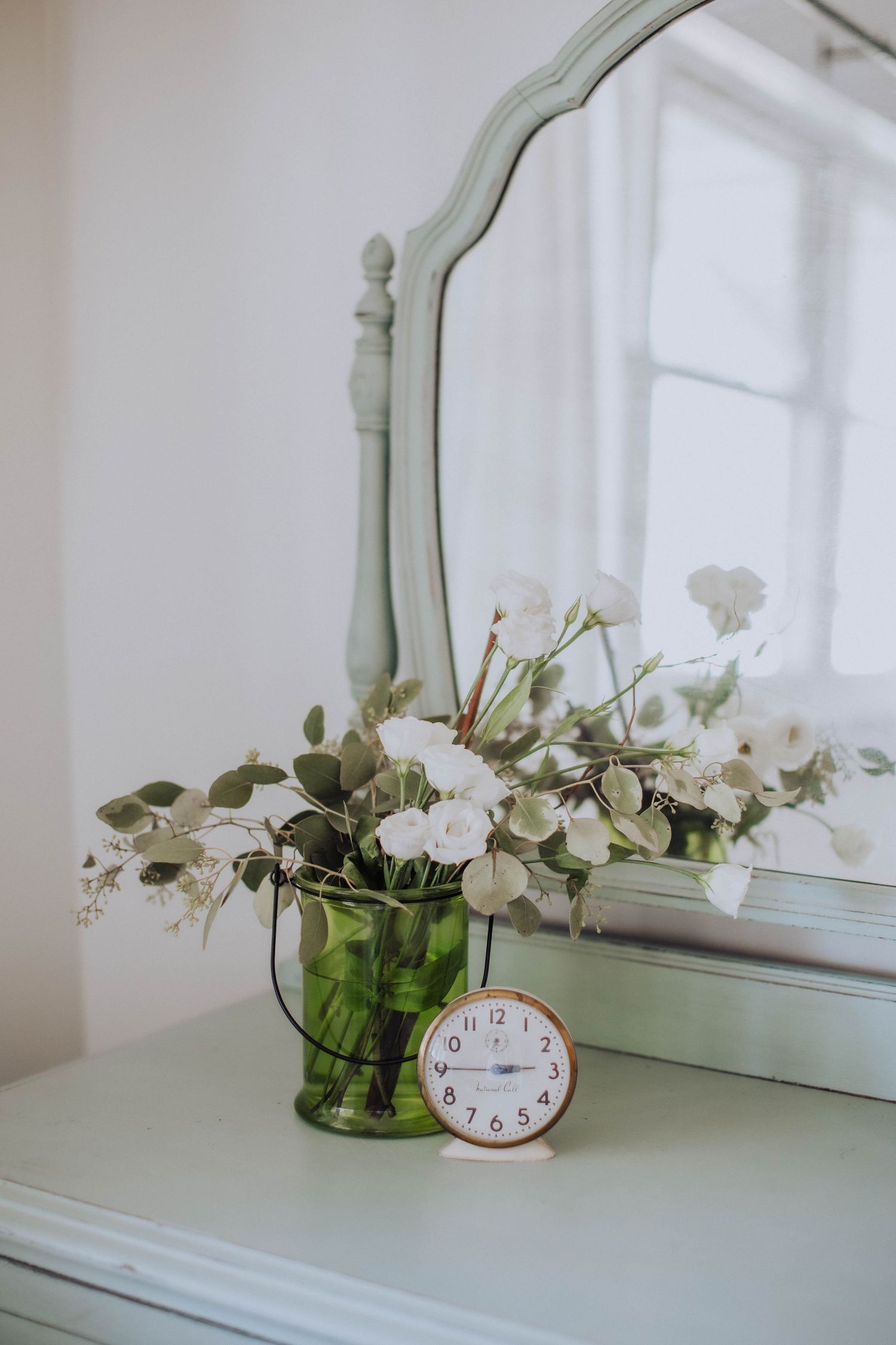 White flowers in a vase next to an analog clock on a mint green dresser top.