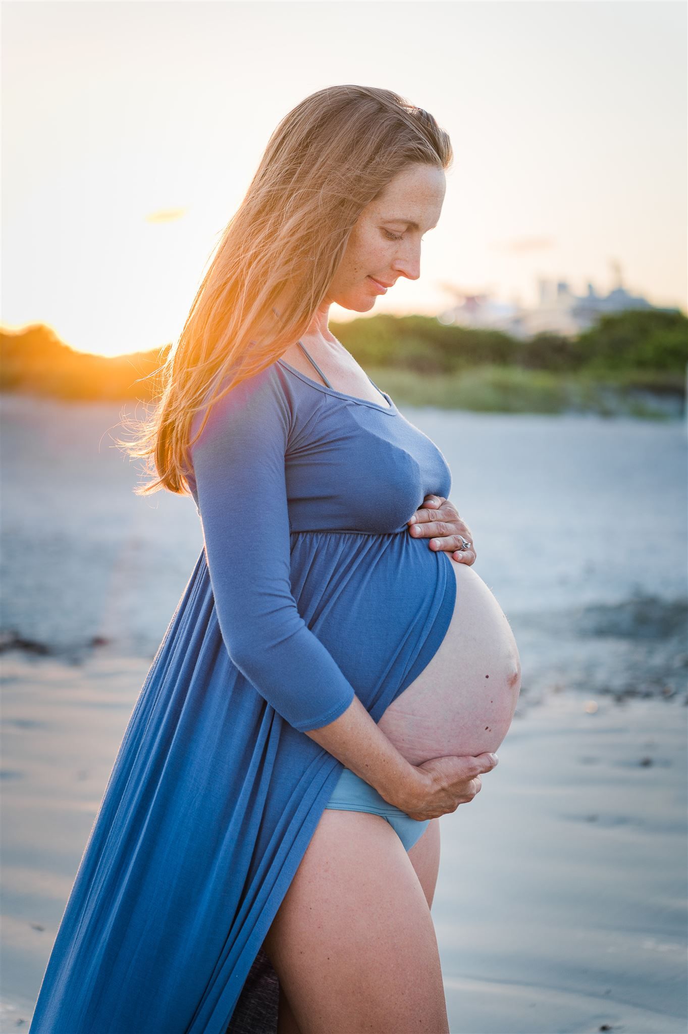 Pregnant woman in blue dress poses contemplatively on beach at sunset.