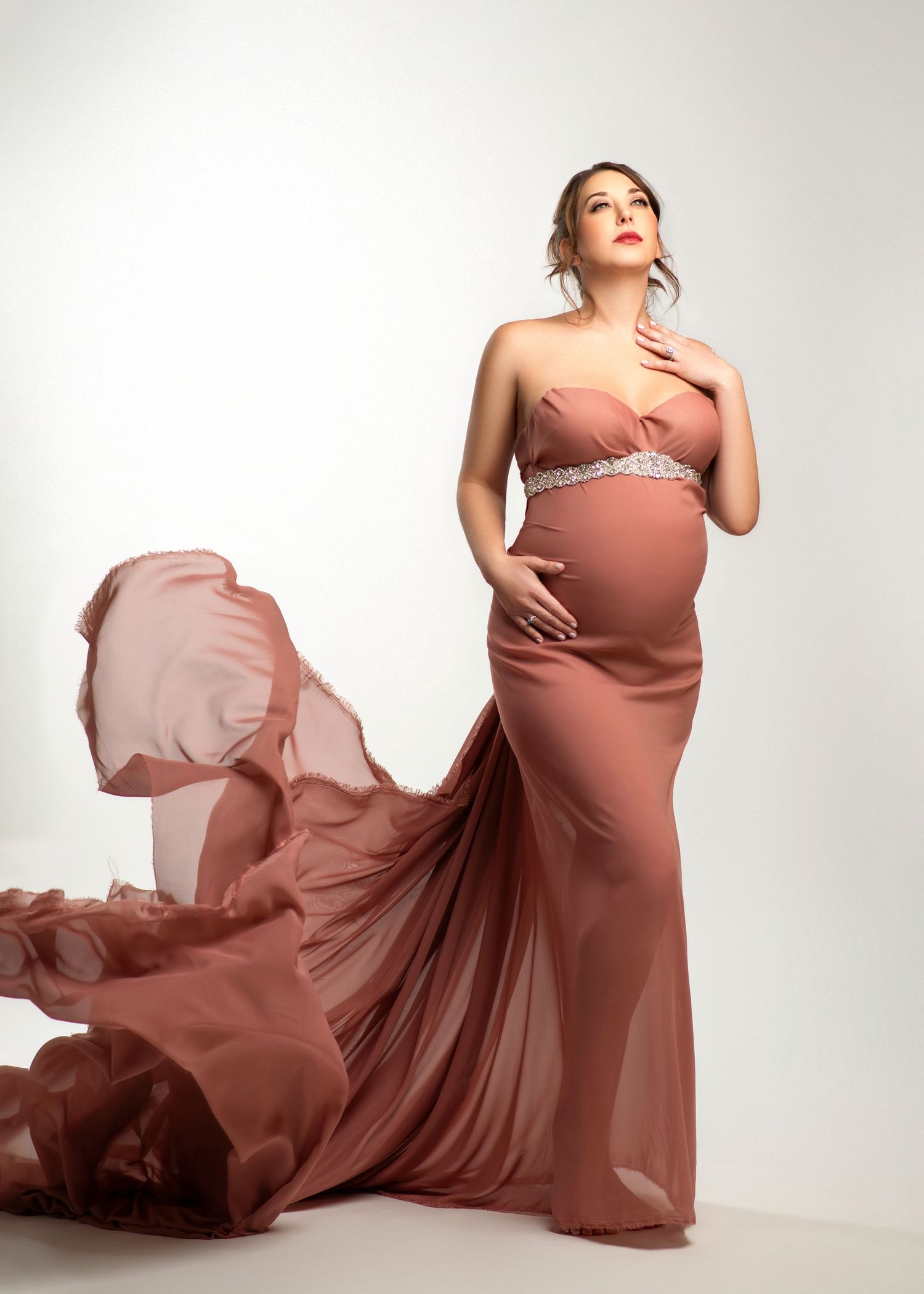 a pregnant woman looks up into the light while wearing a flowing rose gown