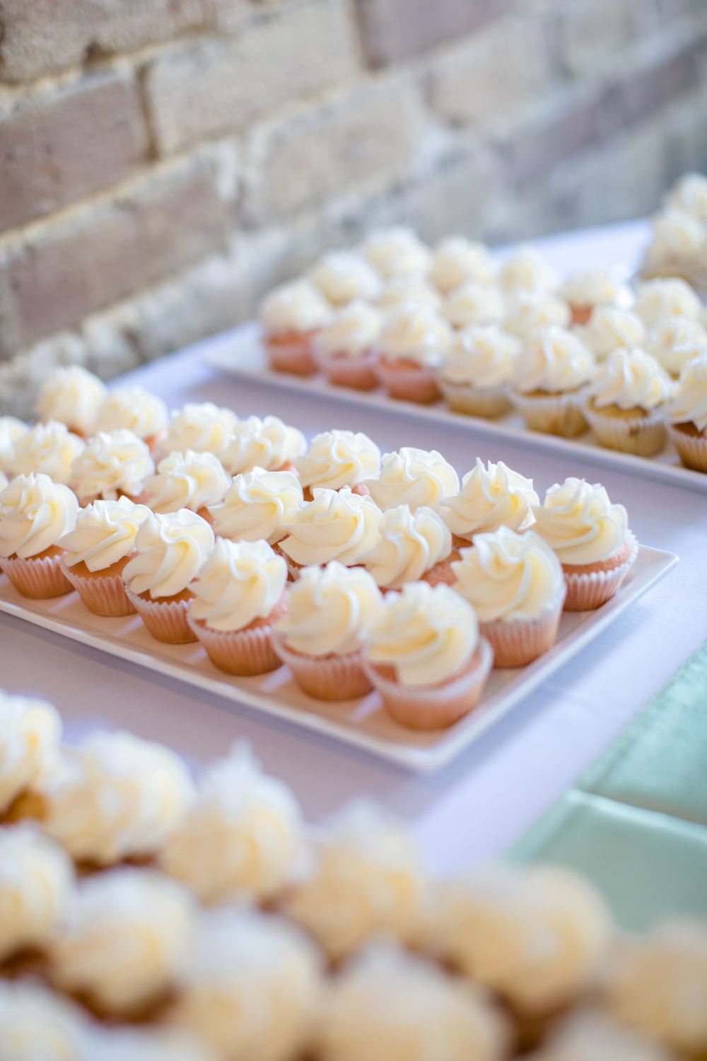 Rows of delicate mini cupcakes with swirled white frosting displayed on white rectangular platters.