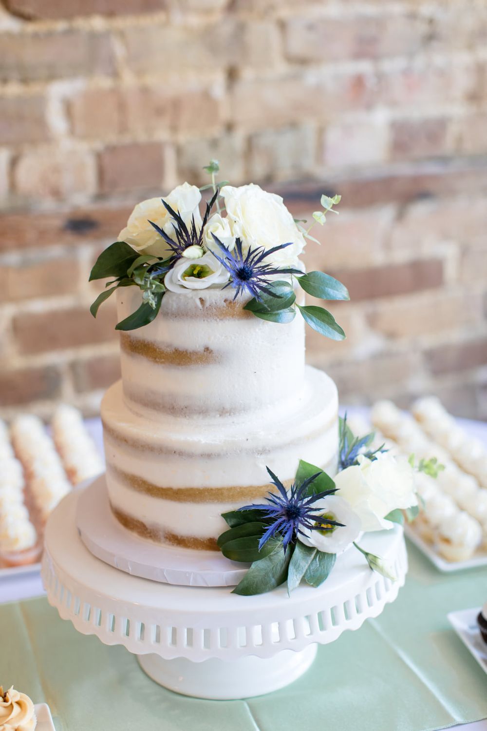 Semi-naked wedding cake decorated with white and blue flowers on a white pedestal stand.