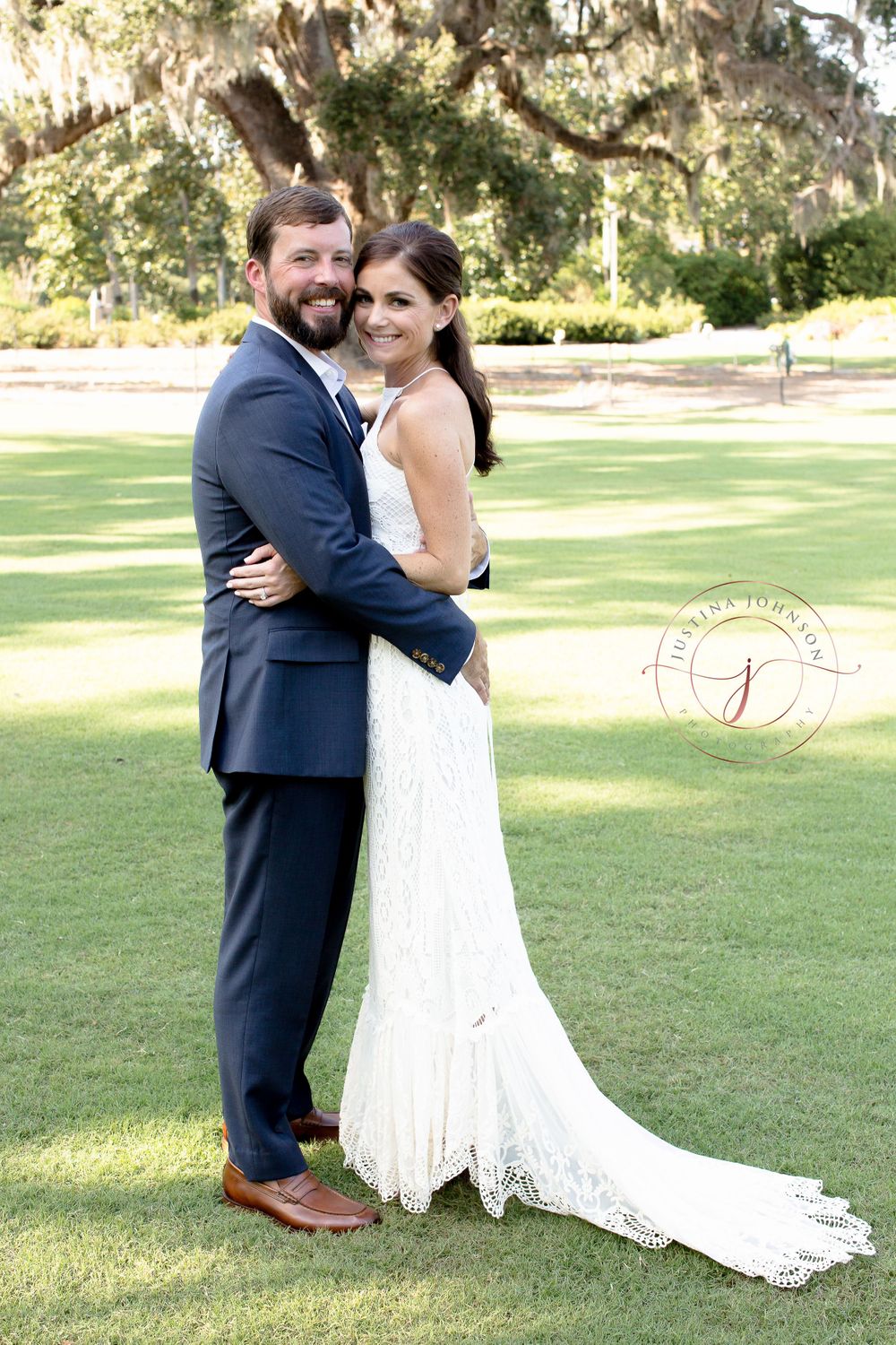 A newlywed couple embraces on a scenic lawn with Spanish moss hanging from trees in the background.