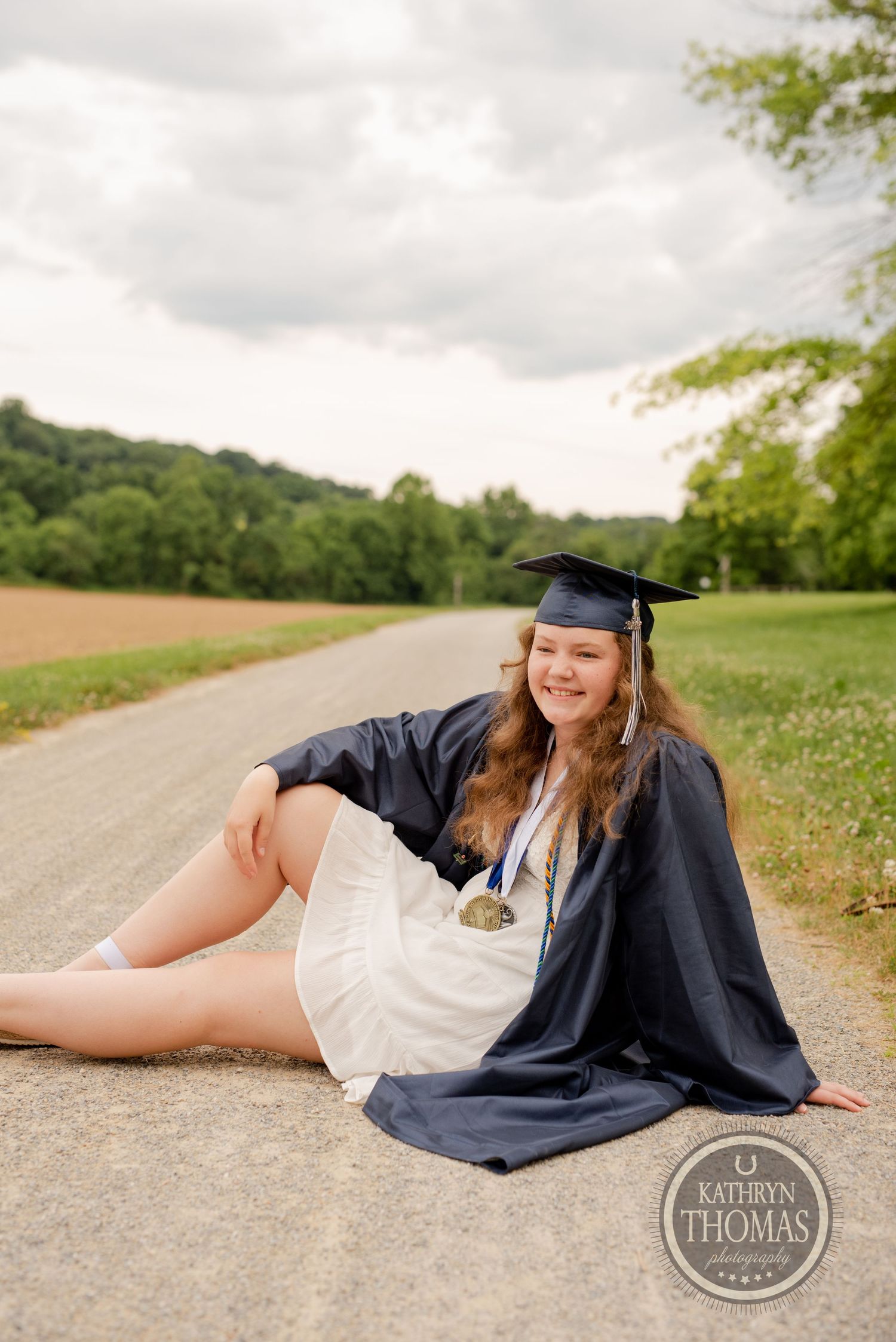 Graduate in cap and gown reclines on rural path during outdoor photo session.