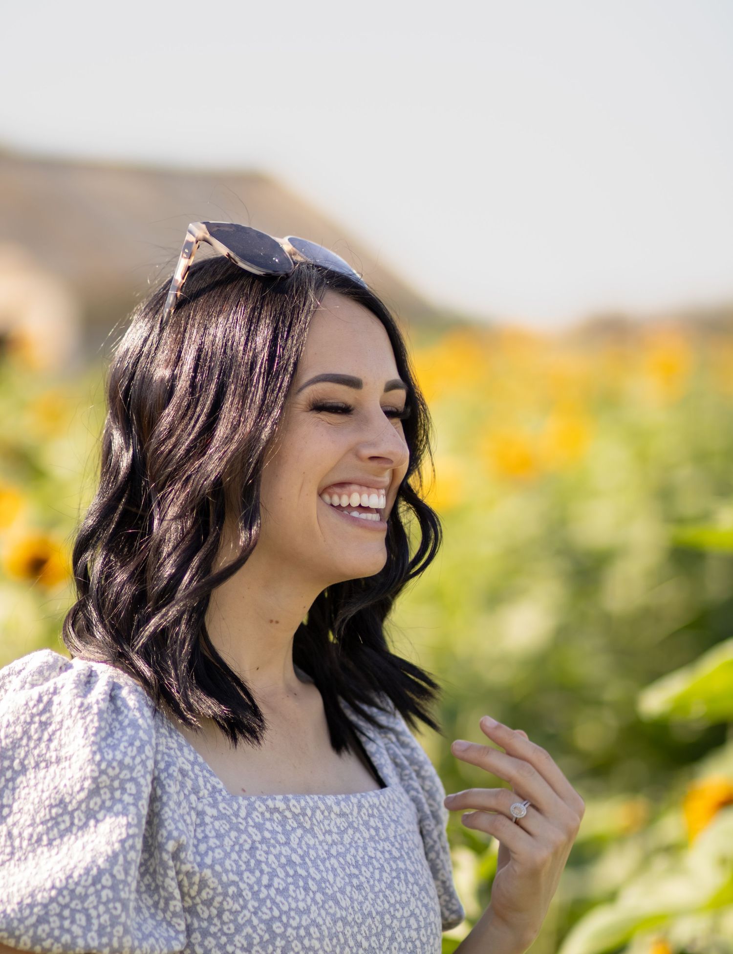 Smiling woman with dark hair and sunglasses in a sunflower field wearing a floral dress on a sunny day.