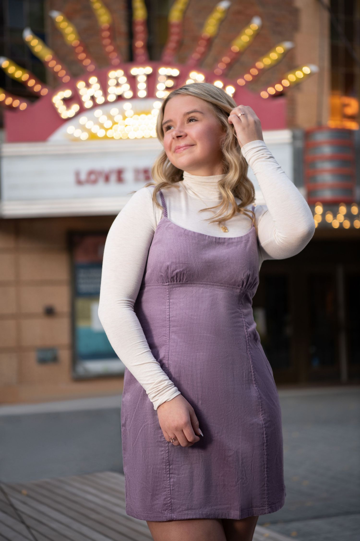 A person in a white turtleneck and purple dress poses in front of illuminated theater marquee lights at dusk.