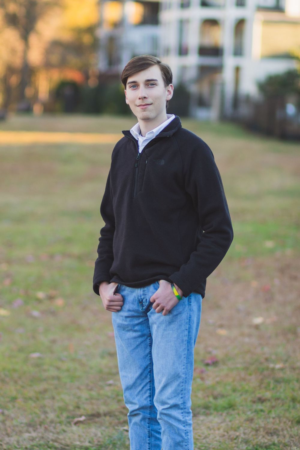 Young person in a dark sweater and jeans poses casually on a grassy area near buildings.