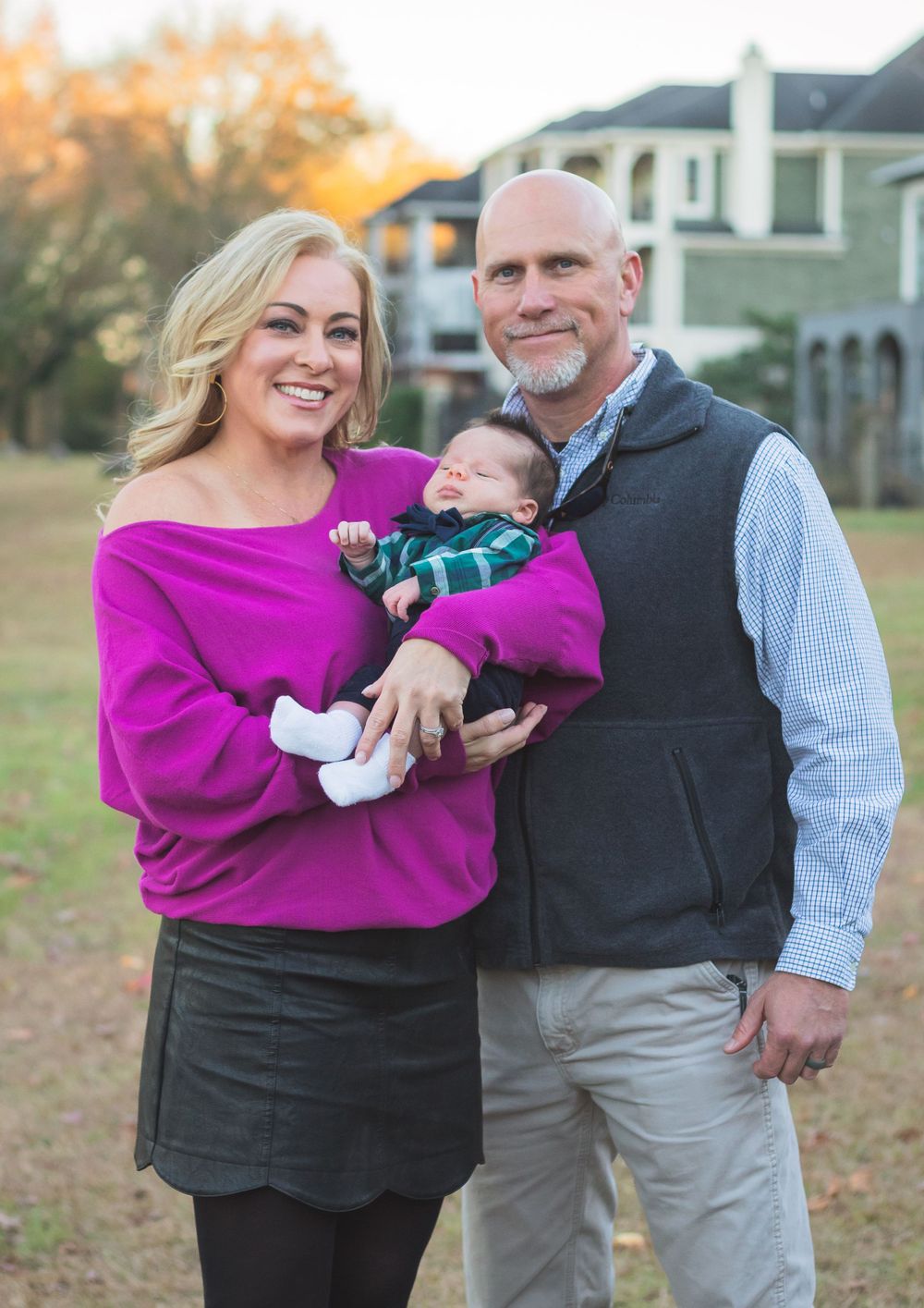 Couple in casual attire holding infant outdoors with residential buildings in background at sunset.