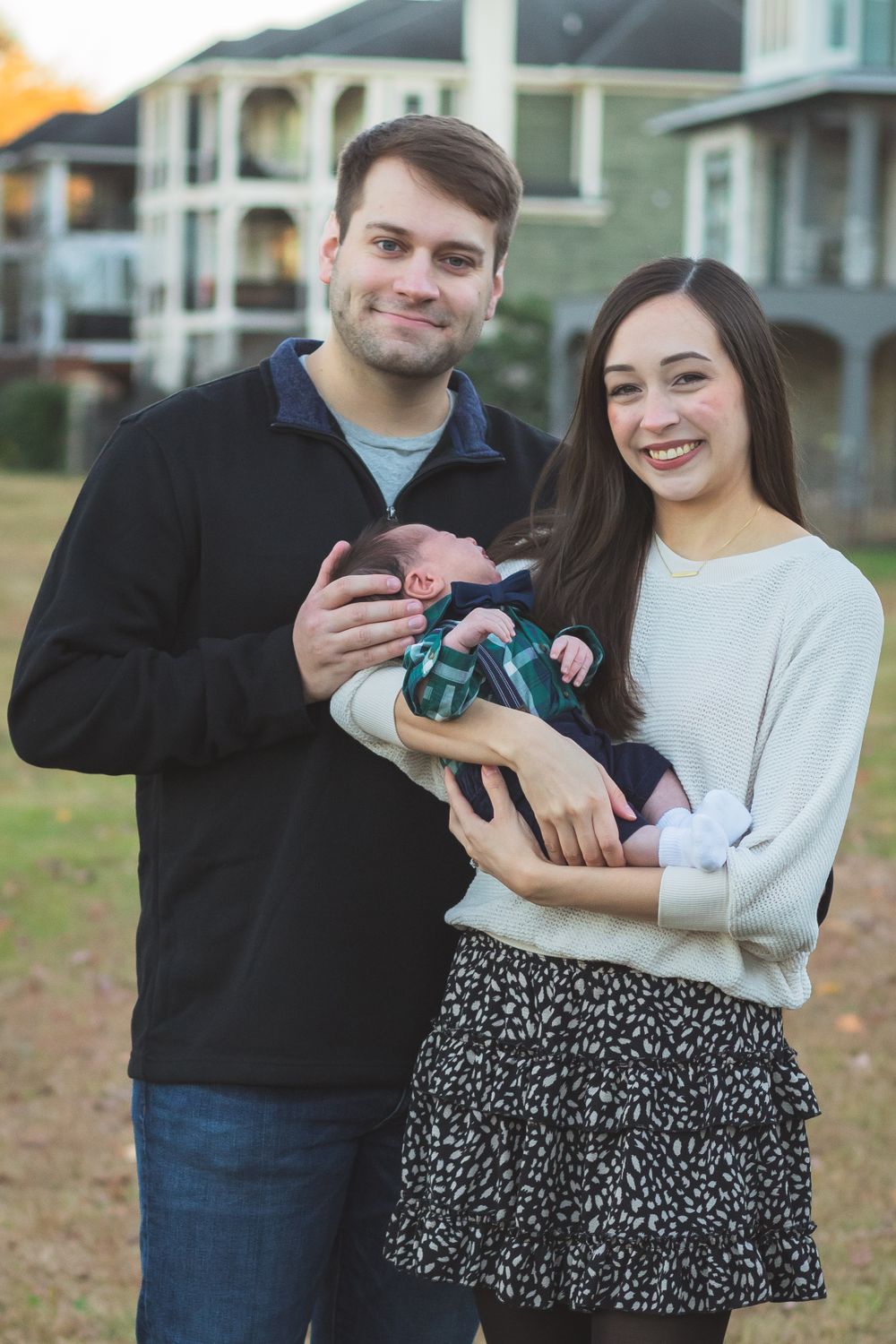 Couple in matching sweaters holding small baby wrapped in blanket outside house.