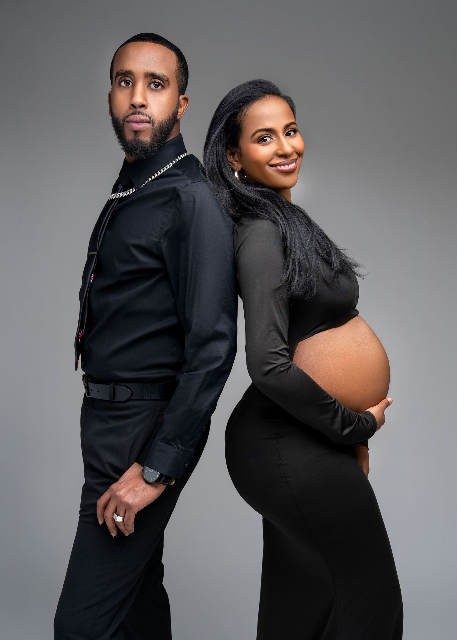 an expecting couple stand back to back smiling