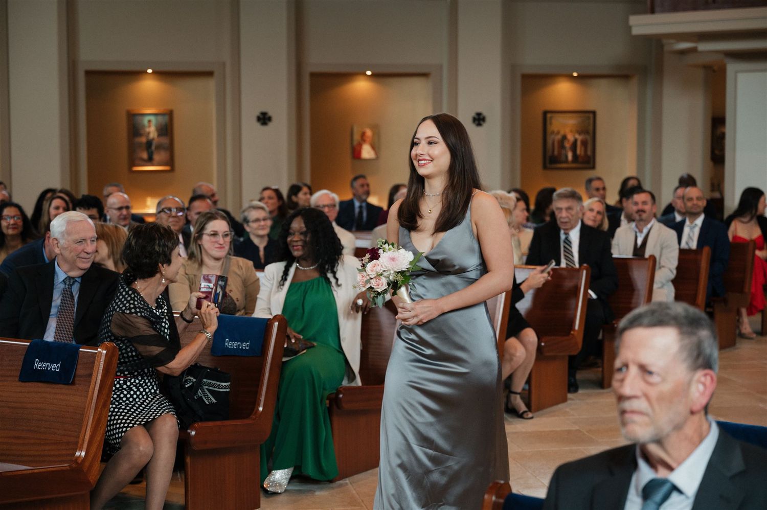 Bridesmaid in silver dress walking down church aisle carrying bouquet.