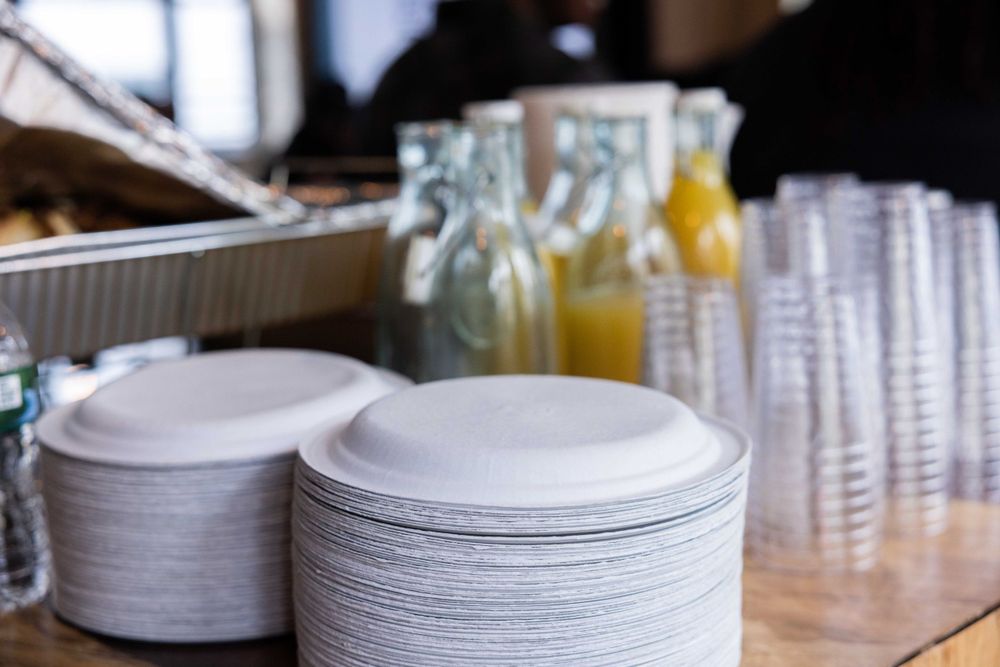 Stacks of white paper plates and clear plastic cups arranged on a counter with bottles in background.