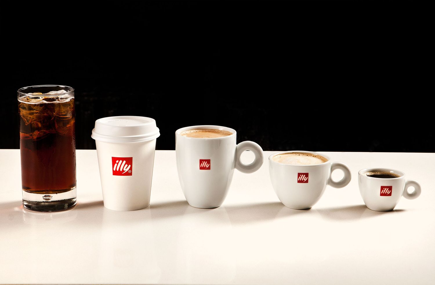 Different sizes of illy coffee cups and glasses arranged in ascending order against a black background.