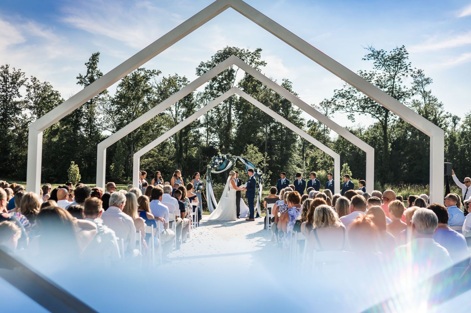 Modern outdoor wedding ceremony under geometric white arches with large crowd.