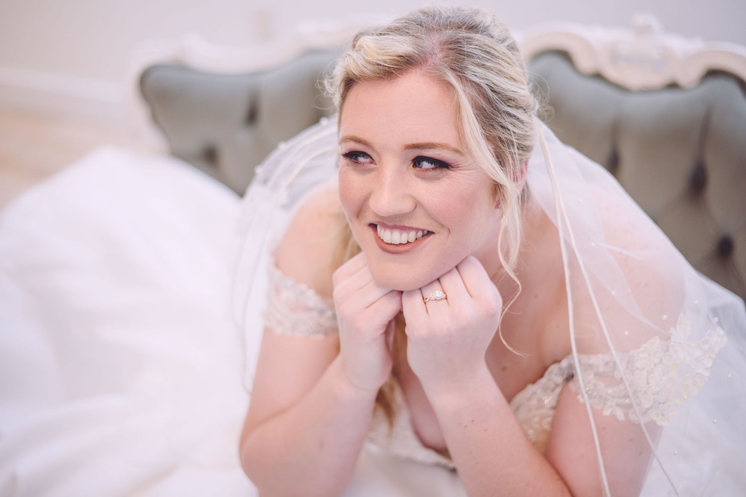 A bride in a white dress and veil reclines on an elegant gray sofa.