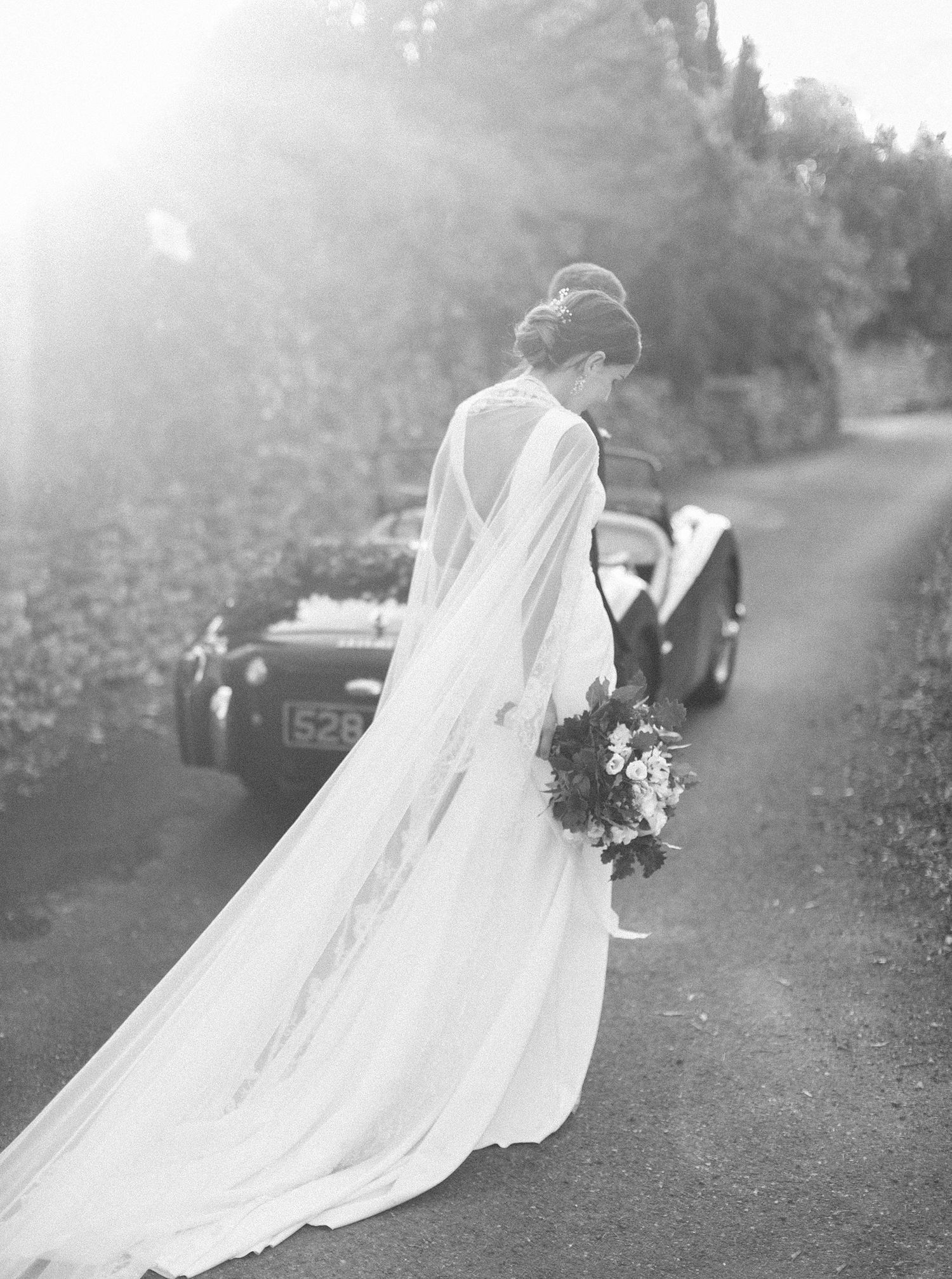 A bride in a long white dress stands gracefully in front of a vintage car at a wedding in Gordes, Provence.