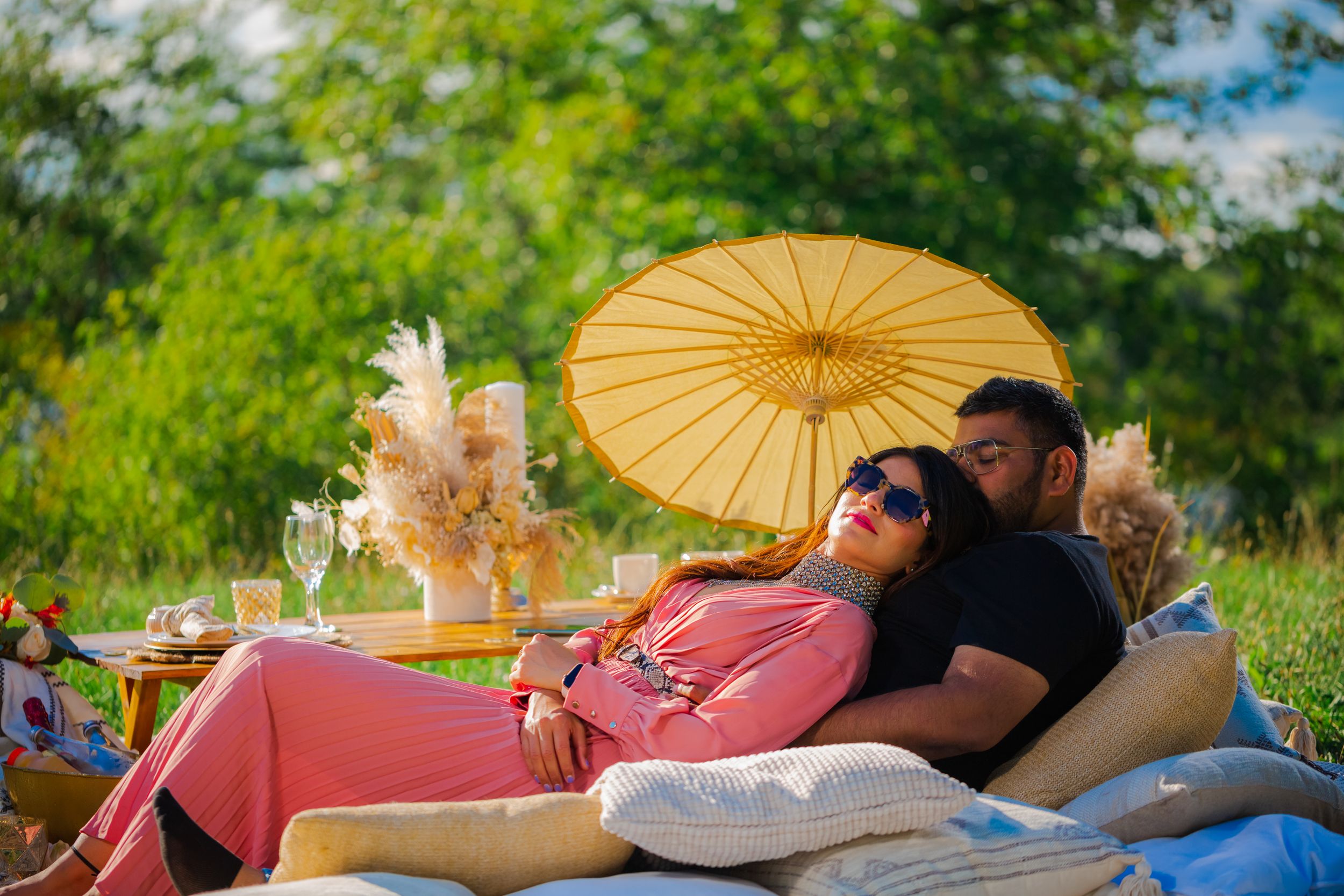 Outdoor picnic setup with blankets and parasol in lush green setting.