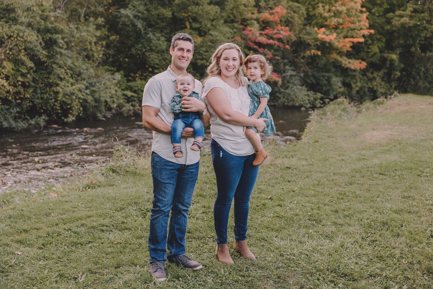 Young family of four poses together on grassy hillside with autumn trees and river in background.