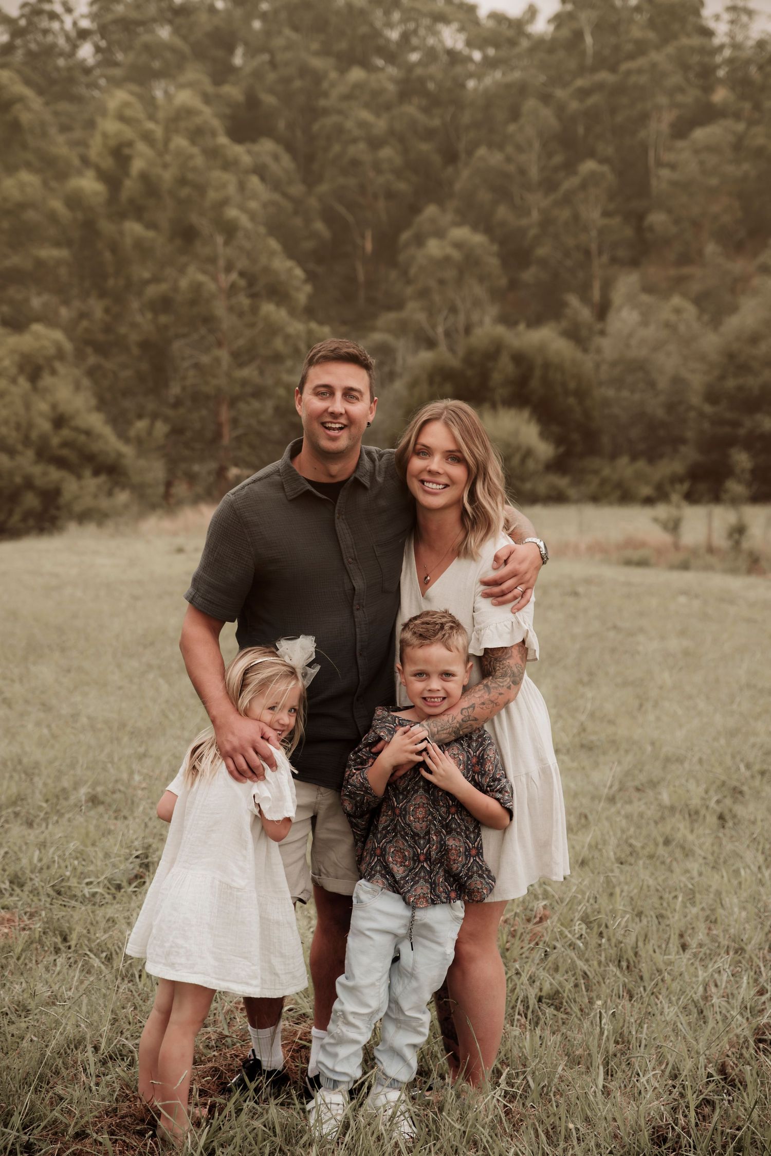Family portrait captured outdoors in a natural field setting with forest backdrop in warm sepia tones.