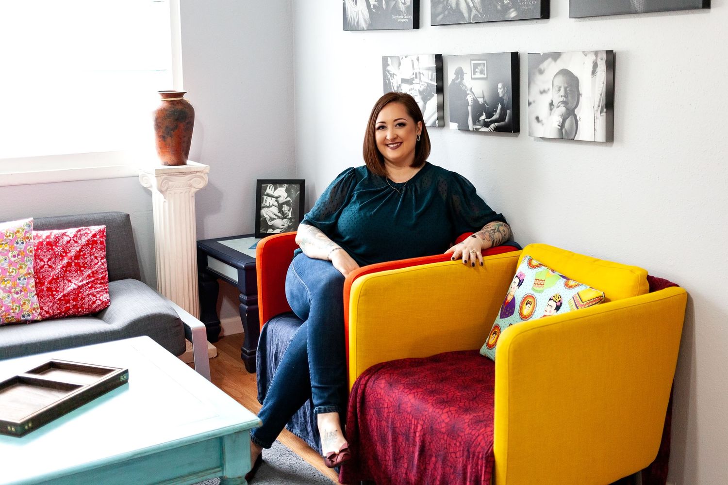 a woman sits cross legged in an orange chair next to an empty yellow chair. A turquoise table is in front of her.