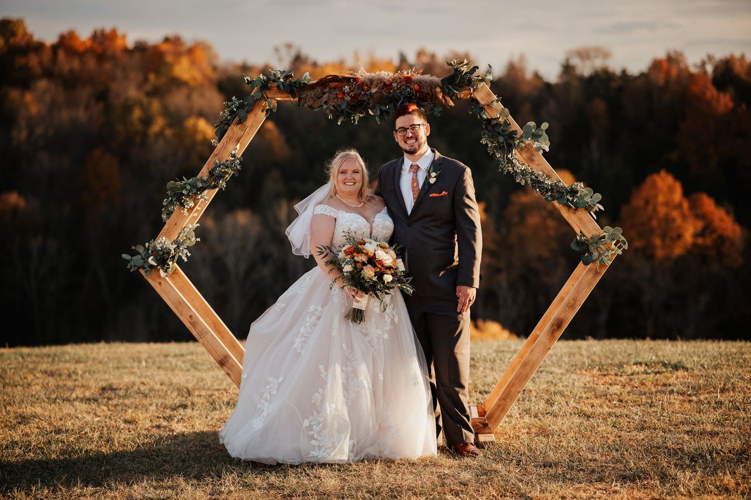 A wedding ceremony under a rustic hexagonal arch decorated with fall foliage and flowers at sunset.