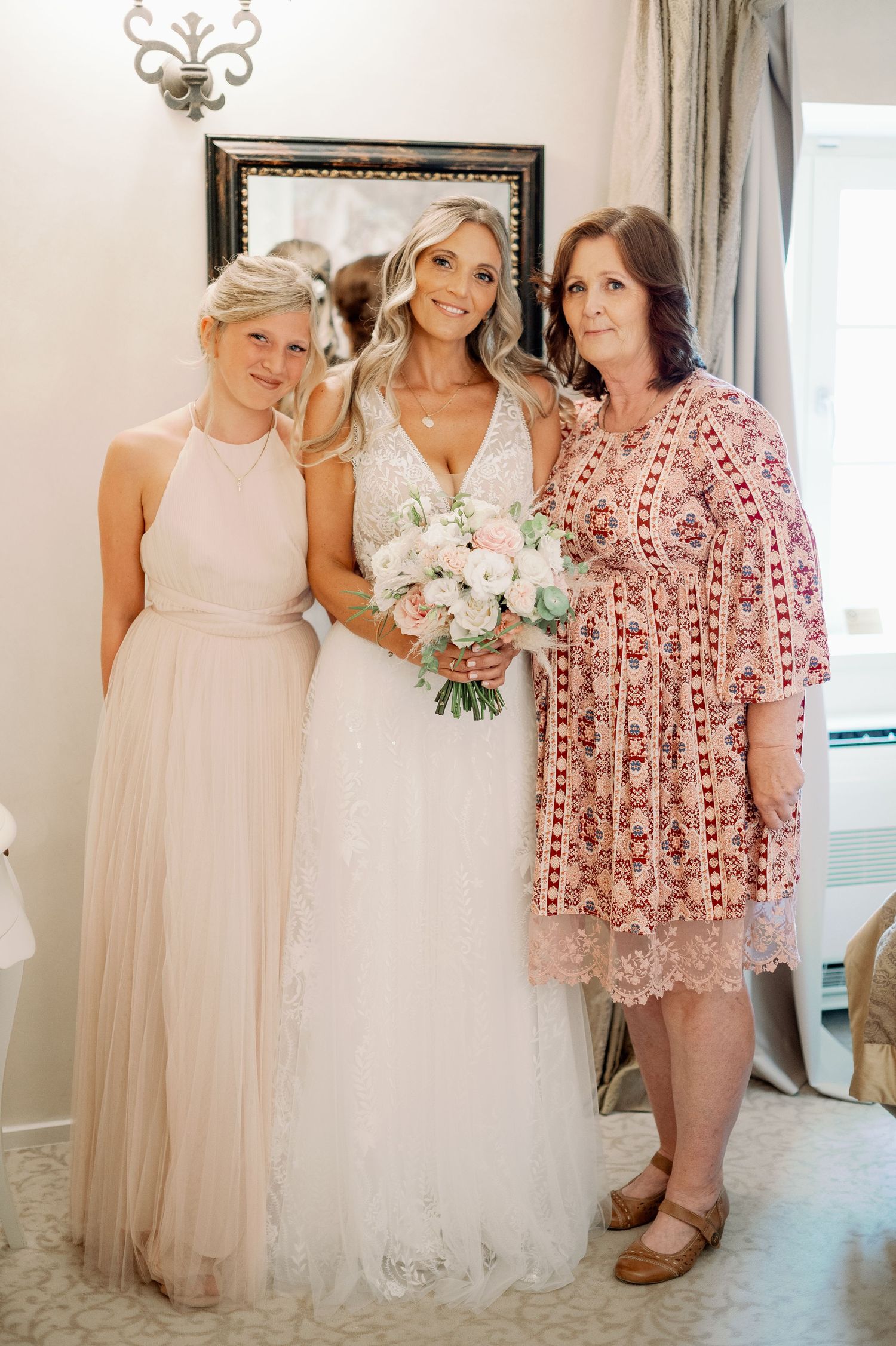 Three women pose together at a wedding, with the bride in white holding a pink and white bouquet.