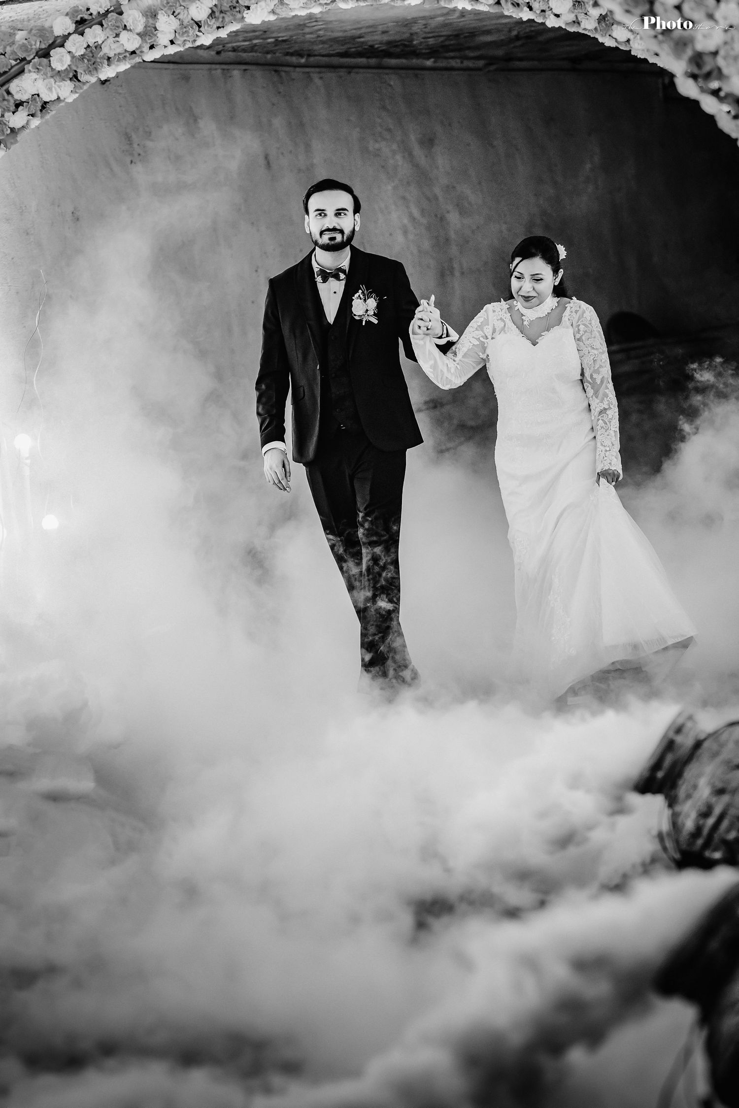 Dramatic black and white wedding photo with couple walking through fog under floral arch.