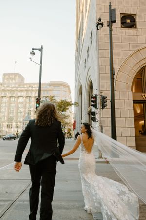 Bride and groom walking hand in hand on city sidewalk beside classic stone building.