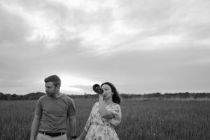 A couple poses in a field during sunset with cloudy skies overhead.