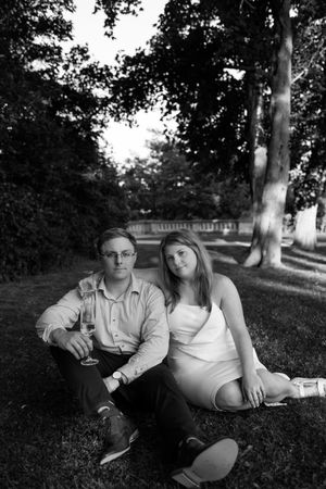A couple sits together on grass in a park setting with trees in the background.