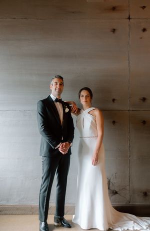 A couple in formal wedding attire poses against a minimal white wall.