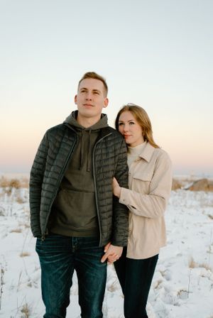Couple in casual winter wear posing together in snowy field at sunset.
