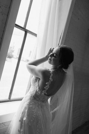 Bride in lace wedding gown poses by window in dramatic black and white portrait.