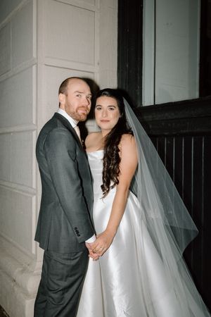 A newlywed couple poses together against a white wall with the bride wearing a long veil.