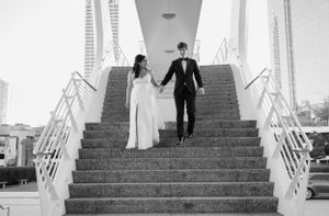 A couple in wedding attire descends white stairs while holding hands.