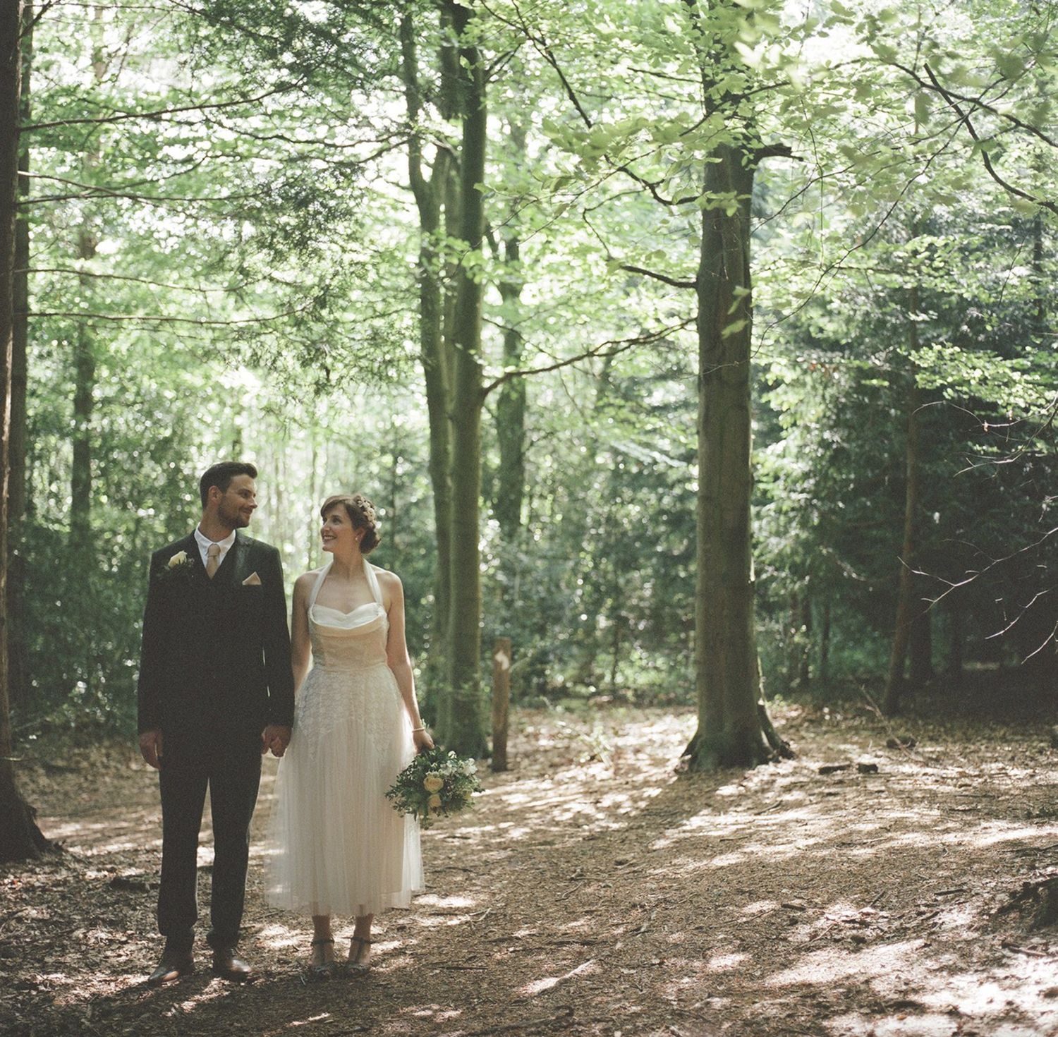 A couple in wedding attire walks through a sunlit forest setting.