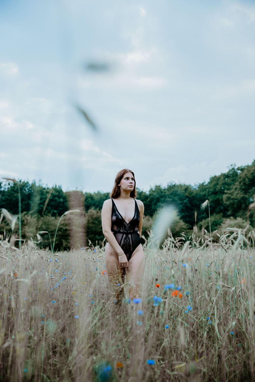 Person poses in field with tall grass and blue wildflowers under cloudy sky.