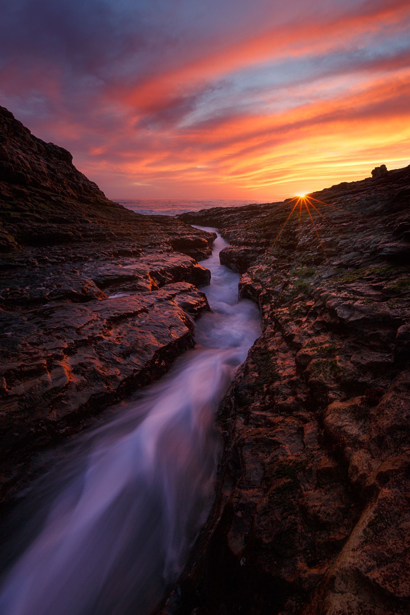 Flowing stream carves through rocky canyon under dramatic orange and purple sunset sky with sunburst on horizon.