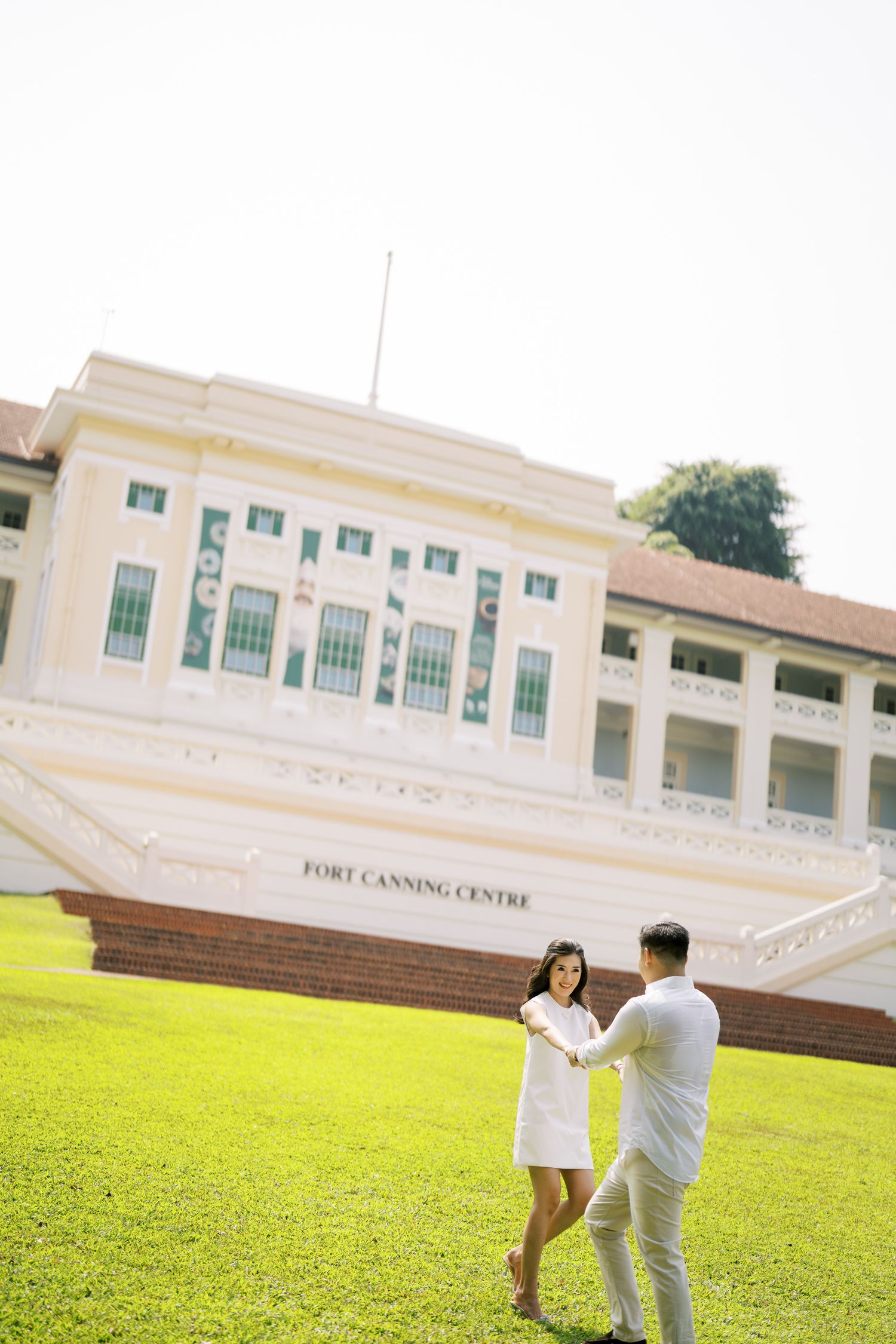 A couple poses playfully on a grassy lawn in front of the Fort Canning Centre building.