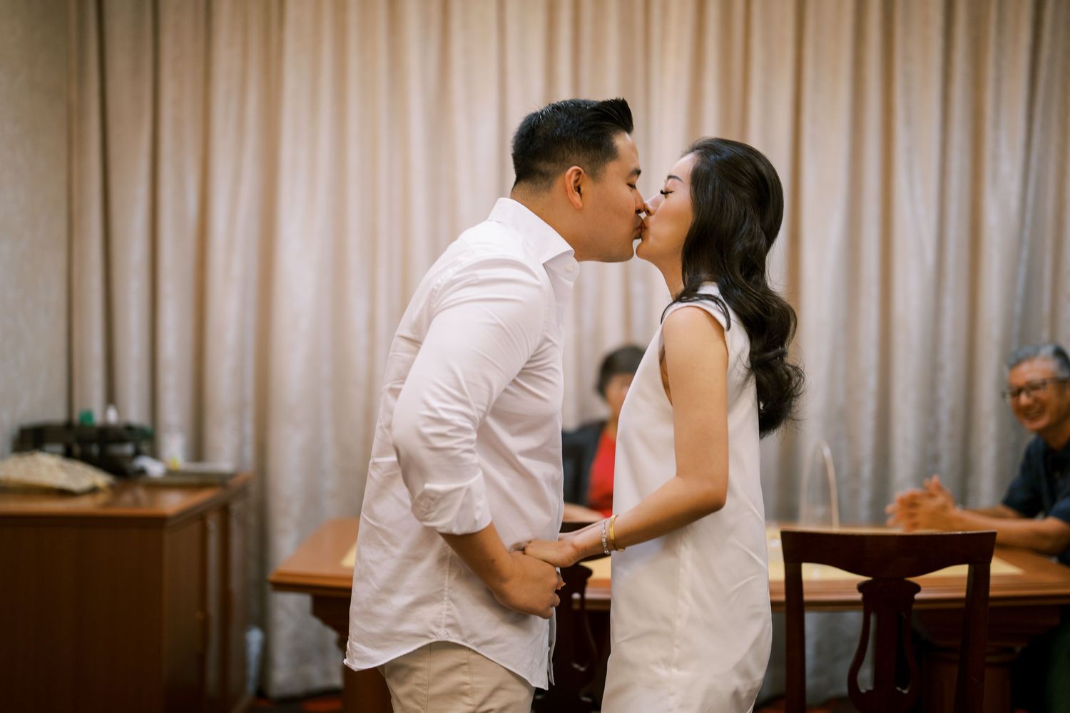 Two people in white clothing sharing an intimate moment in a meeting room setting.