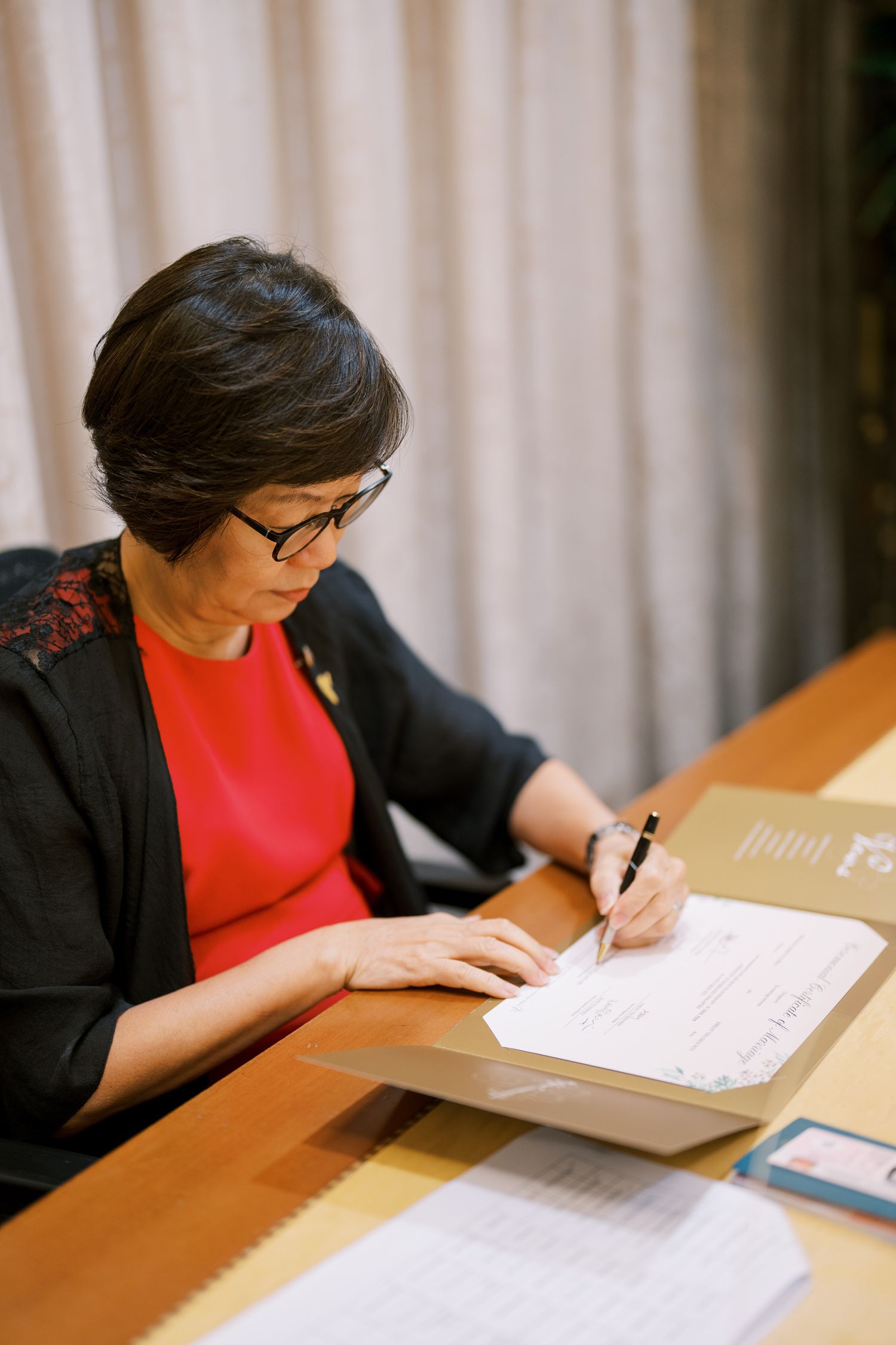 Person in a red top and black jacket writing on documents at a wooden desk.