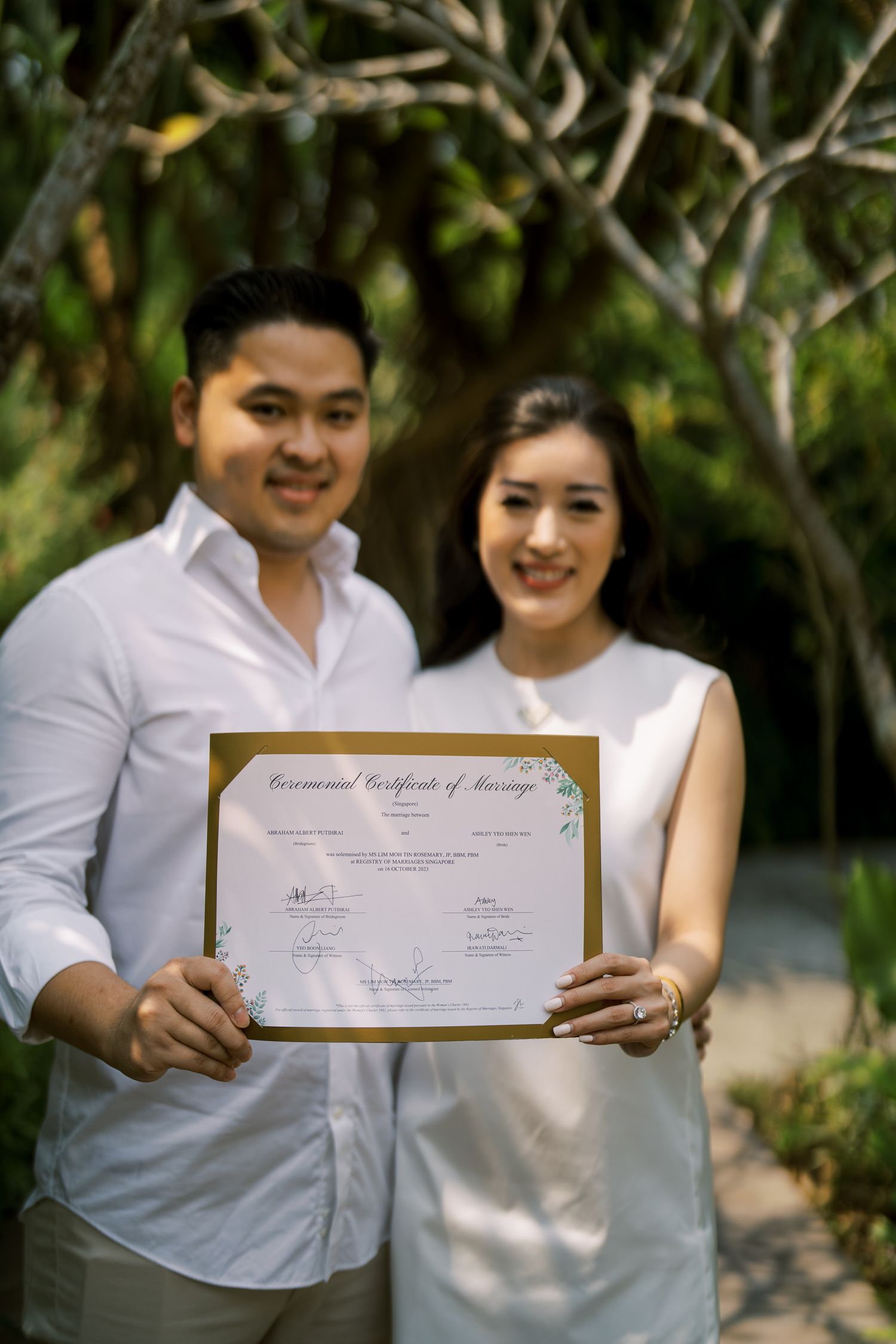 A couple dressed in white holding up a certificate together in a garden setting.