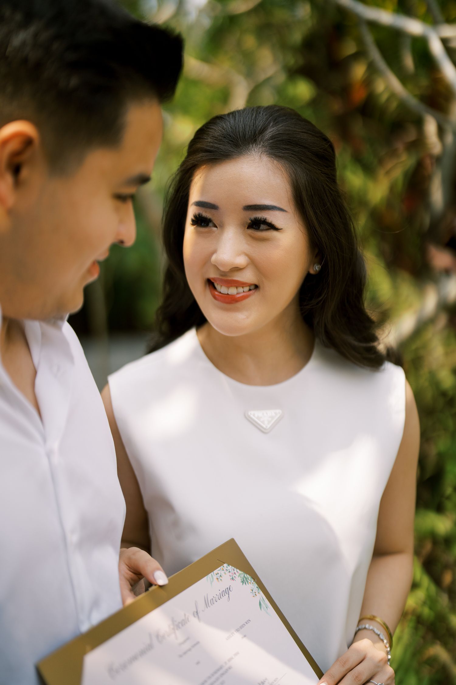 A couple in white outfits sharing smiles while looking at a document in a garden.