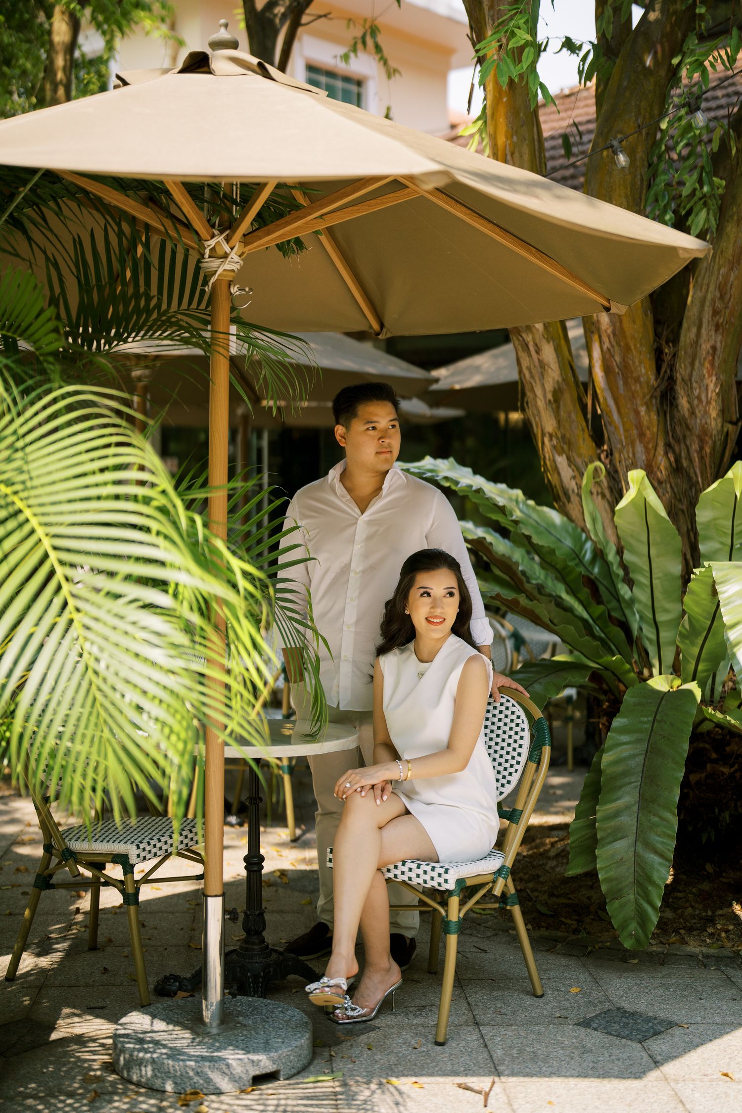 Couple sitting together under beige umbrellas in a tropical garden setting with lush palm fronds.