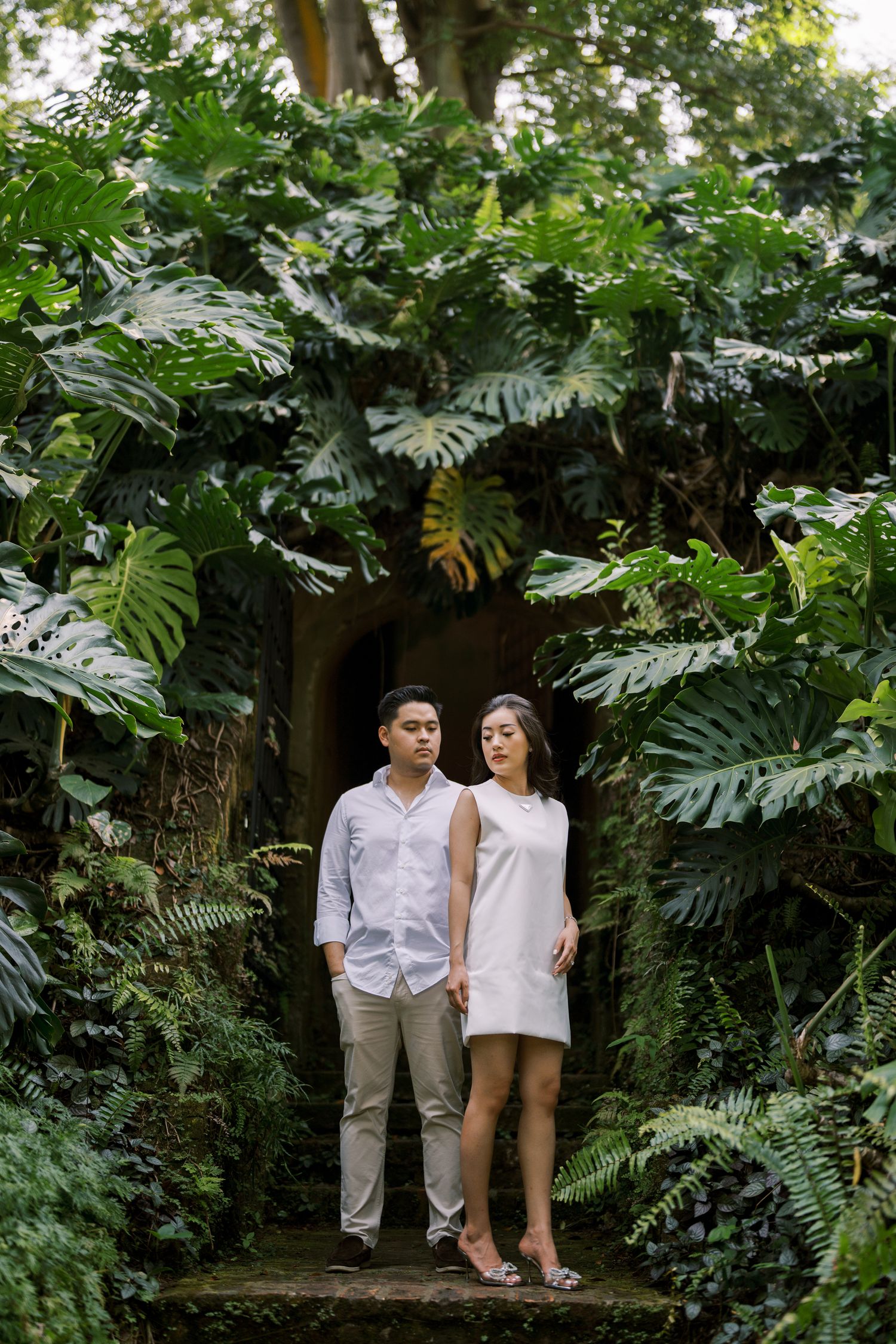 A couple poses together in a lush tropical garden setting with large monstera leaves in the background.