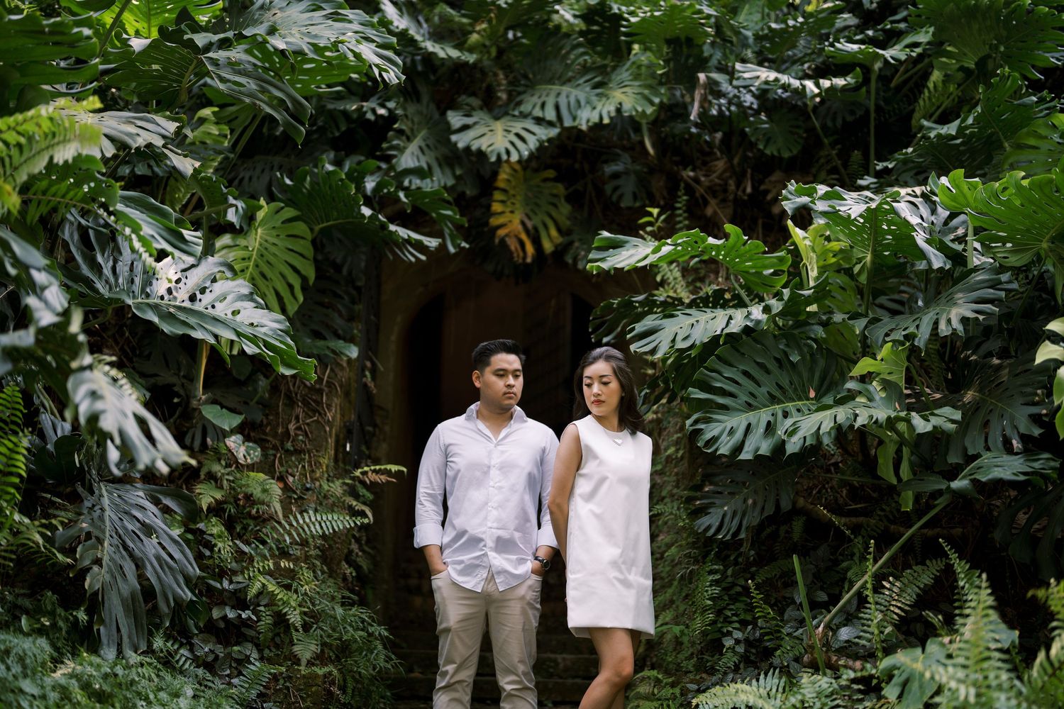 A romantic couple stands together surrounded by vibrant green tropical foliage.