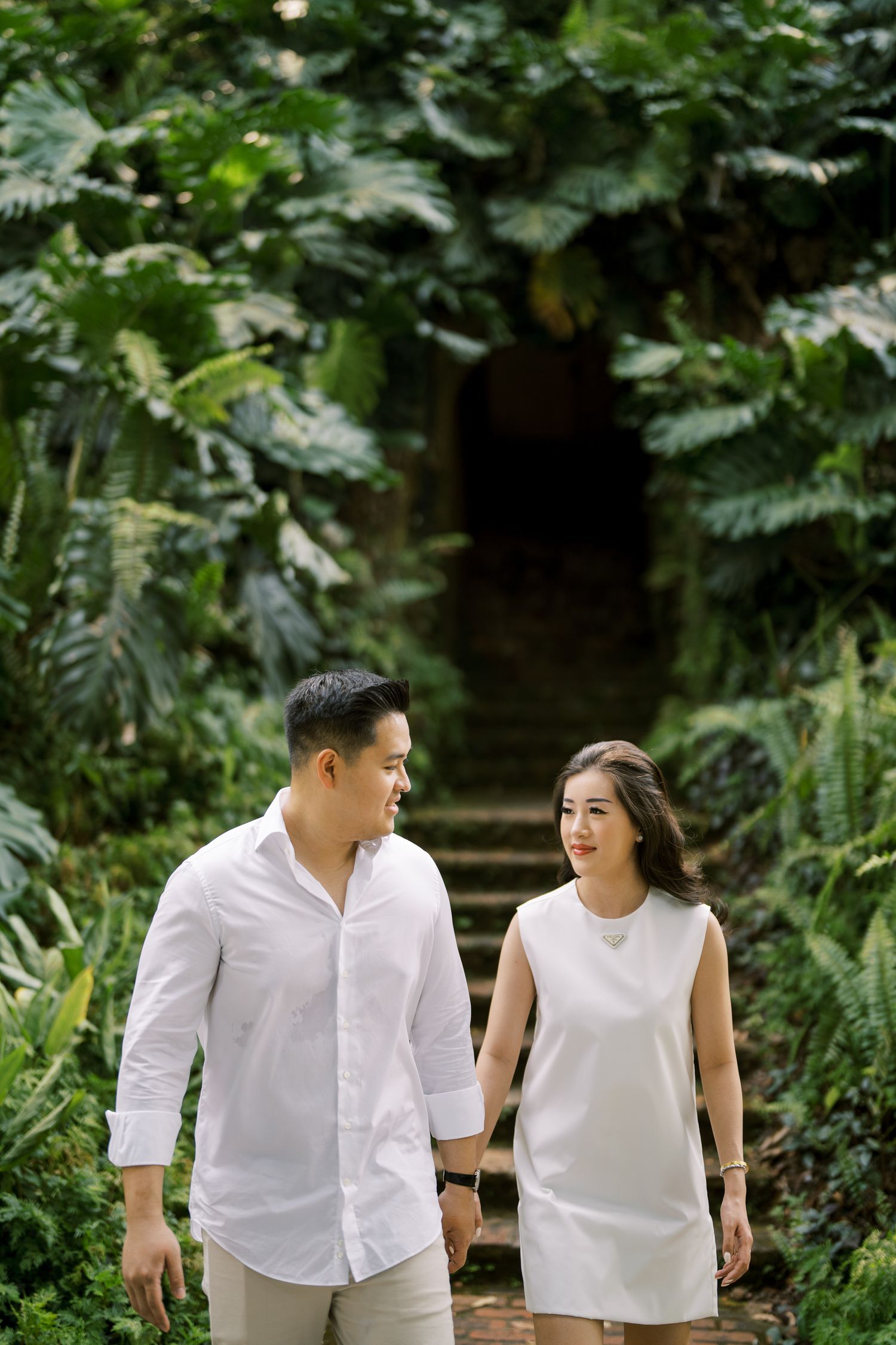 A couple wearing white outfits shares a joyful moment while walking through a garden path.